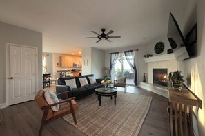 A living room with a sofa, armchair, coffee table, fireplace, and a view of the kitchen and outdoor area.