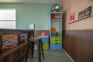 Una habitación con un banco de madera, un taburete, un ventilador y estantes con diversos objetos, incluyendo un cartel que dice 'Mejor Cerveza'.