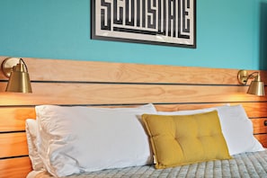 Une chambre à coucher avec une tête de lit en bois, deux lampes de chevet, un lit avec des coussins blancs et moutarde, et une œuvre d’art accrochée au mur.