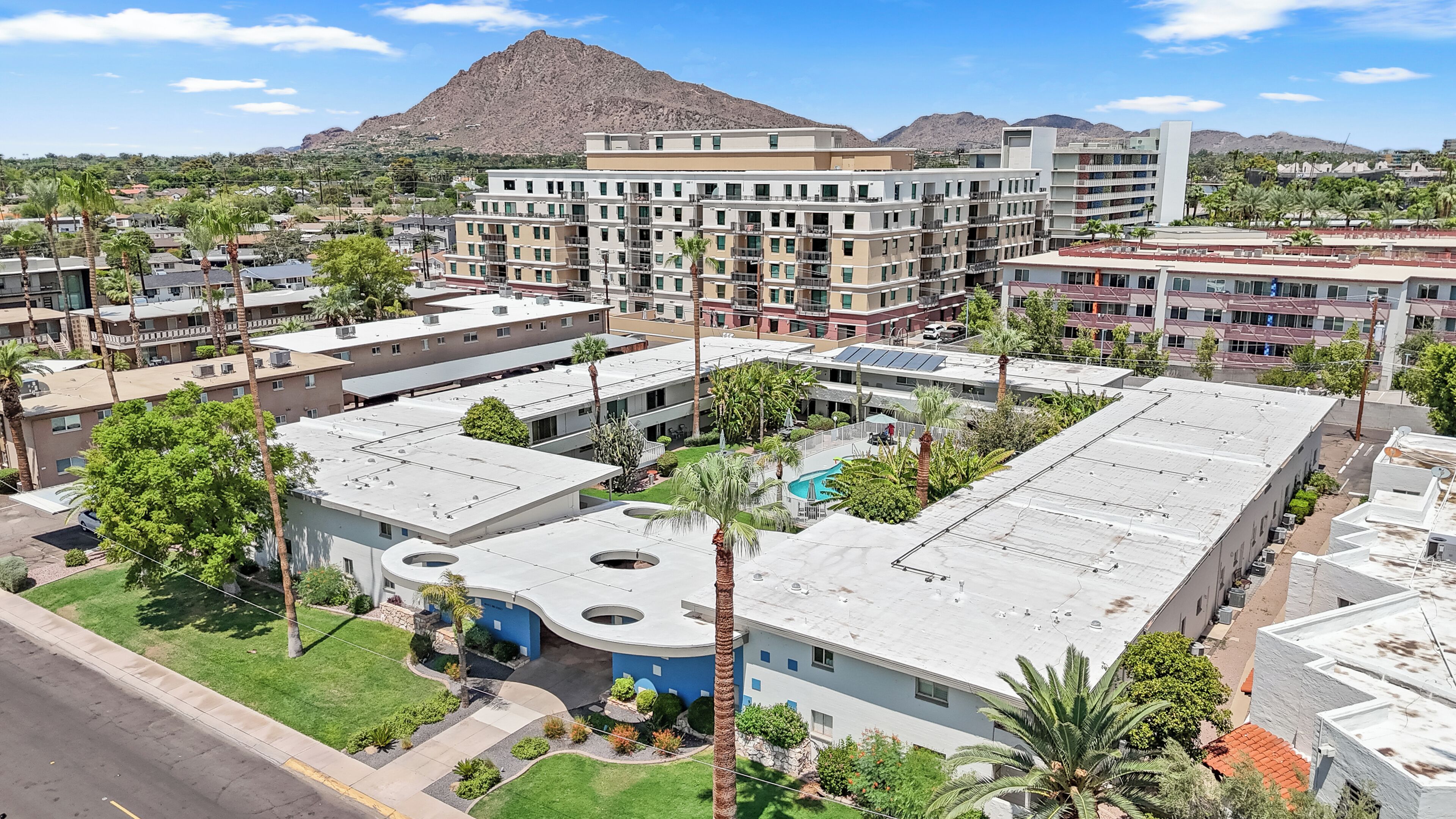 A multi-story hotel with a pool, surrounded by palm trees and a mountainous backdrop.