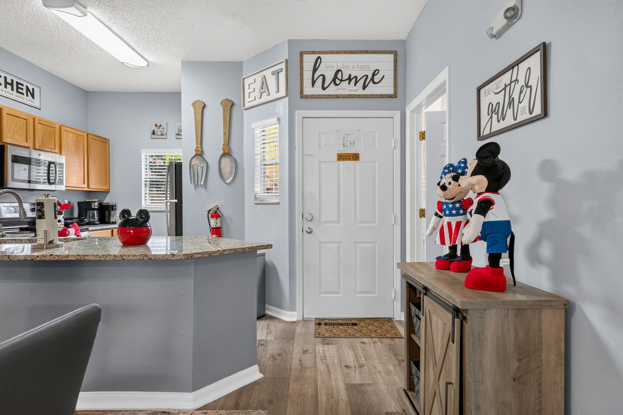 Una cocina con isla central, armarios de madera y un microondas. Hay letreros decorativos en las paredes y una puerta con un cartel encima.