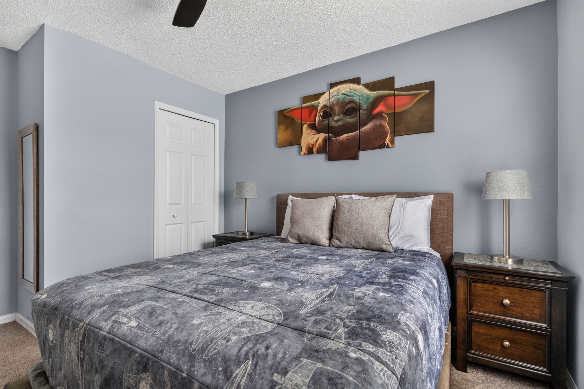 A bedroom with a bed, a nightstand, a lamp, a door, and a wall with a large artwork.