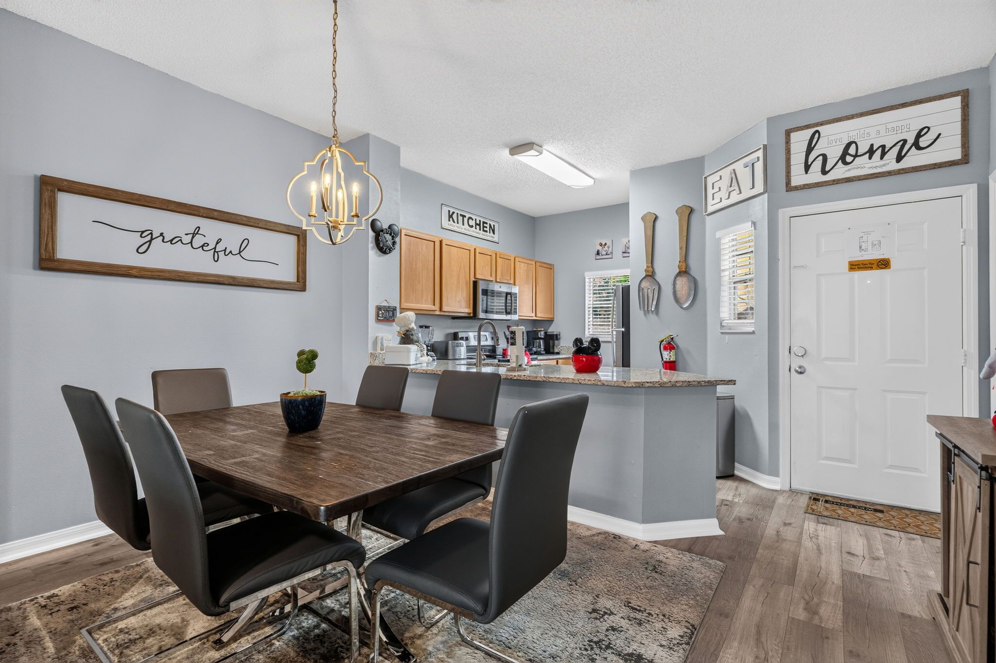 A modern kitchen with a dining area, featuring a chandelier, wooden cabinets, and a kitchen sign.
