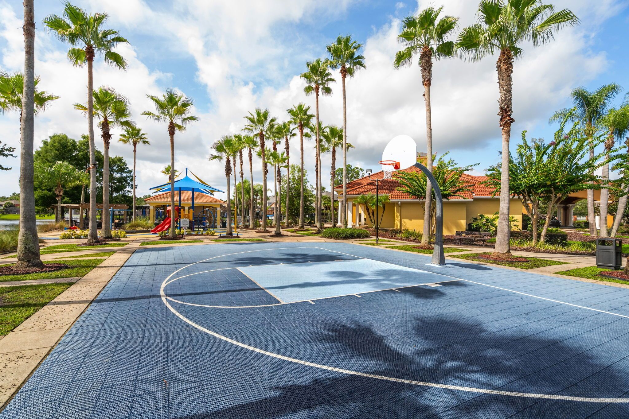 An outdoor basketball court surrounded by palm trees and a playground with a blue roof.