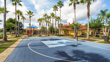 An outdoor basketball court surrounded by palm trees and a playground with a blue roof.