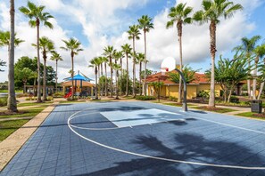 Ein Basketballplatz im Freien, umgeben von Palmen und einem Spielplatz mit blauem Dach.