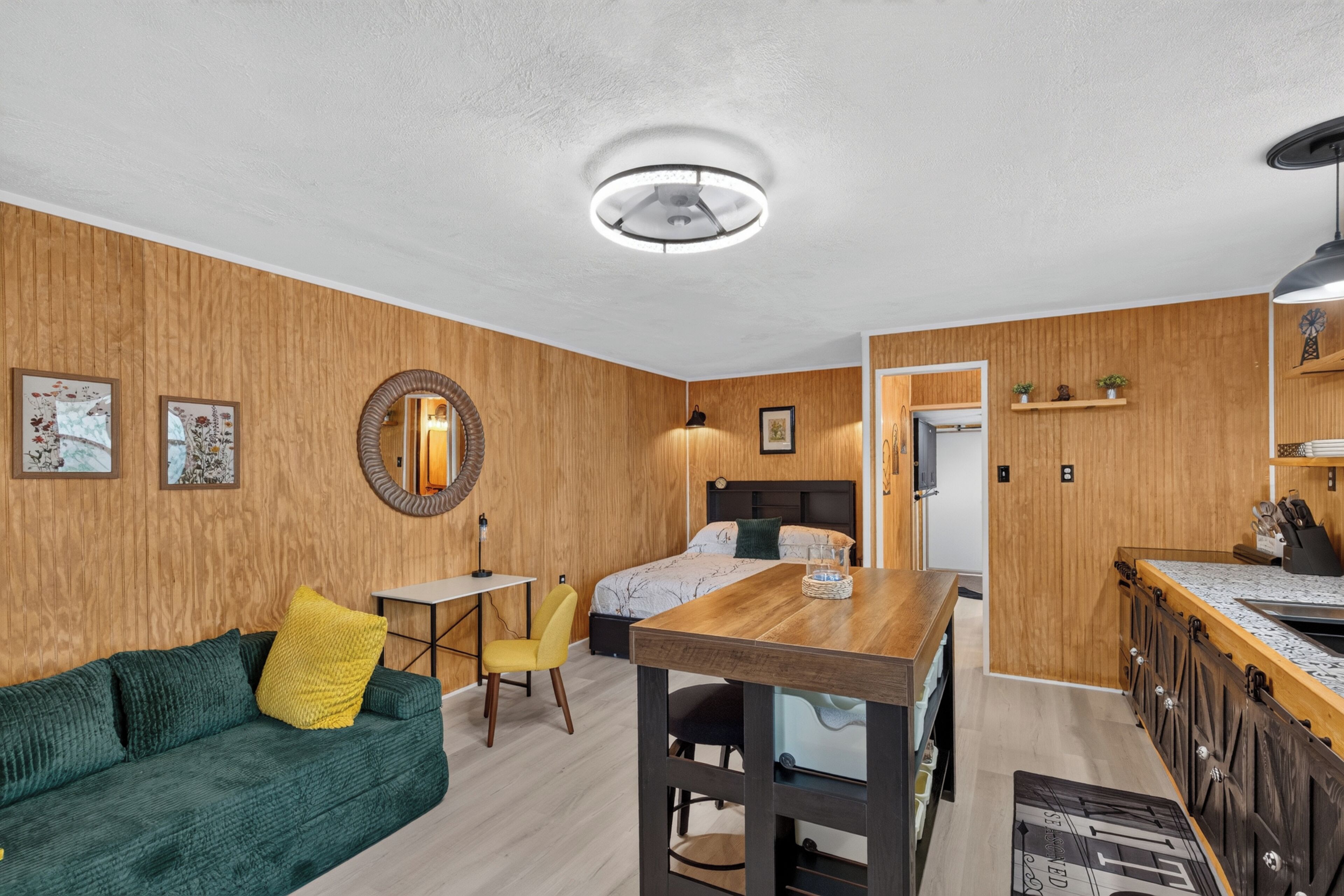 A compact room with a wooden wall, a bed, a desk, a chair, and a small kitchenette.
