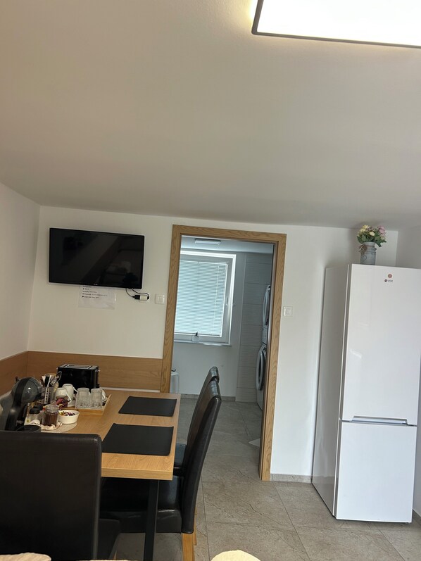 A compact kitchen with a dining area, a flat-screen TV mounted above the counter, and a refrigerator to the right.
