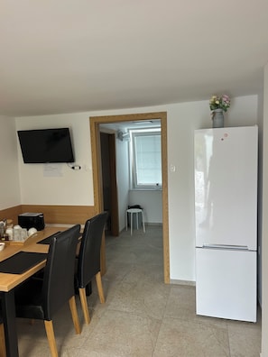 Ett kompakt kök med en platt-TV, ett vitt kylskåp, ett matbord med stolar och en liten pall.