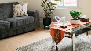 A living room with a sofa, a glass coffee table, a potted plant, and a window with blinds. - Entire Westminster Townhouse Near WestminsterAbbey (London)