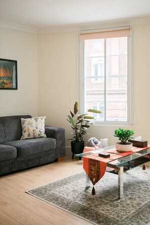 A living room with a sofa, a glass coffee table, a potted plant, and a window with blinds. - Entire Westminster Townhouse Near WestminsterAbbey (London)