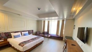 A hotel room with a large bed, a TV, a seating area, and a window with curtains.
