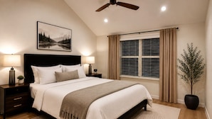 Quarto com cama, mesas de cabeceira, ventilador de teto, janela com cortinas e um quadro pendurado na parede.