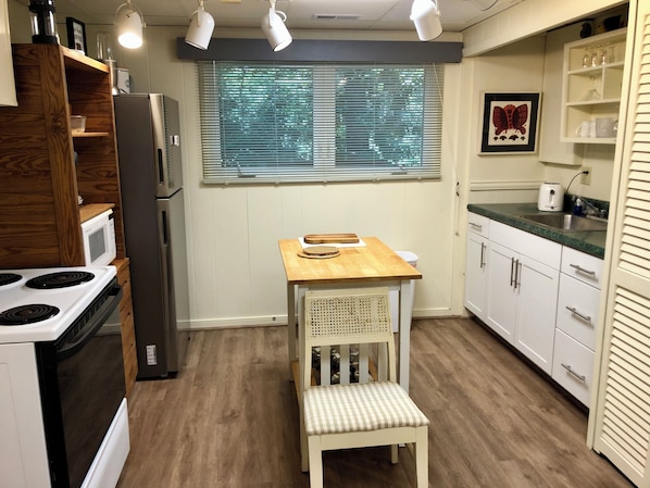 A compact kitchen with a wooden table, a built-in bench, a refrigerator, a microwave, and a window with blinds.
