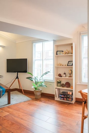 A living room with a flat-screen TV, a bookshelf with decorative items, a potted plant, and a wooden floor. - Two Bedrooms Flat Near Big Ben & Westminster Abbey (London)