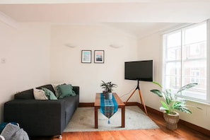 A living room with a dark-colored sofa, a flat-screen TV on a tripod stand, a small table with a plant, and a window with a view of buildings. - Two Bedrooms Flat Near Big Ben & Westminster Abbey (London)