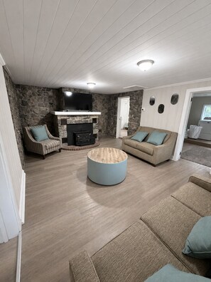 A living room with a stone wall, a fireplace, a television, and a round coffee table. - The Stone House • 5 Min to JMU • Fenced Yard • Peaceful Retreat No Neighbors (Harrisonburg)