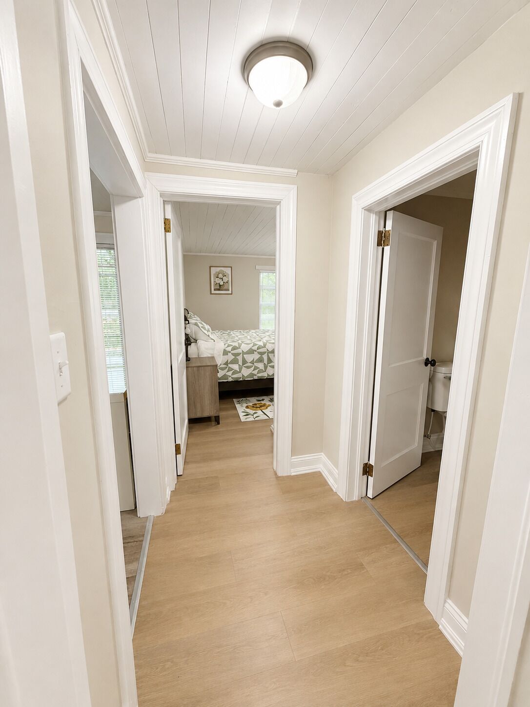 A hallway with a wooden floor leading to a bedroom with a bed and a window.