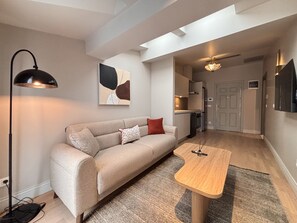 Ein modernes Wohnzimmer mit einer Couch, einem Couchtisch und einer Lampe. An der Wand hängt ein Gemälde und eine Tür führt zur Küche.