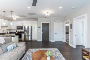 A modern kitchen with stainless steel appliances, a dining area with a sofa and a coffee table, and a hallway leading to a door.