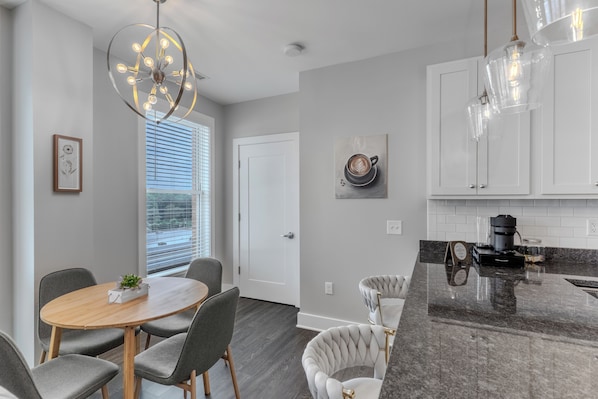 A modern kitchen with a marble countertop, a dining area with a round table and chairs, and a view through the window.
