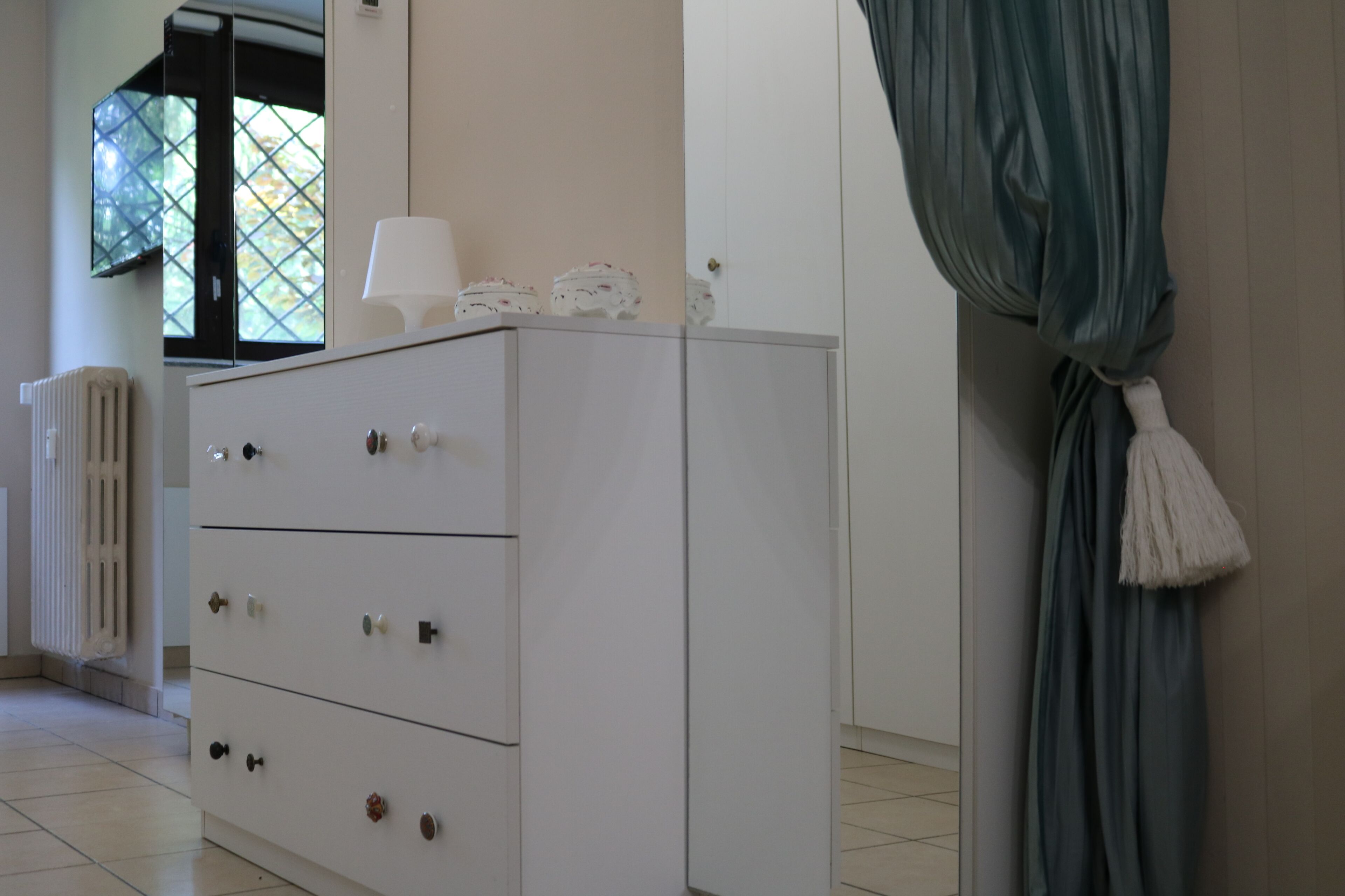 A white dresser with drawers and knobs, a radiator, a window with a view, and a curtain.