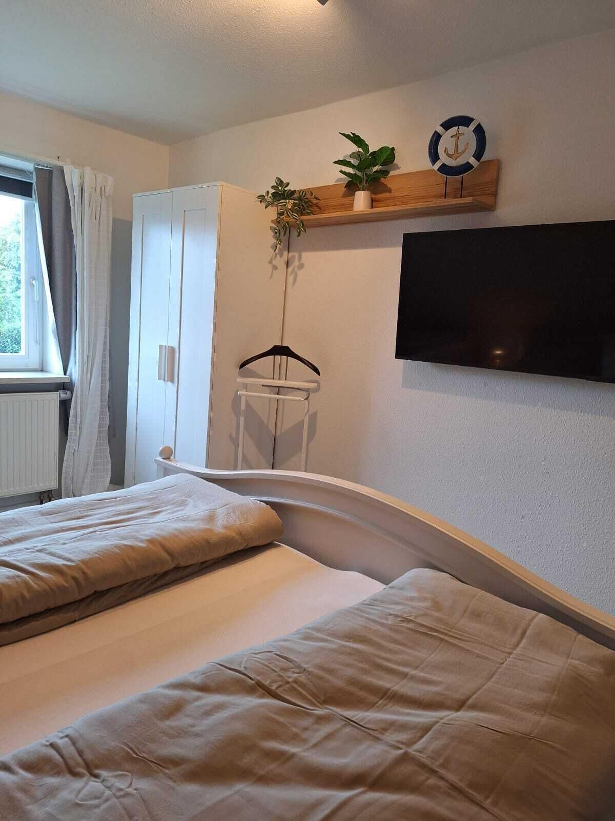 Un dormitorio con dos camas, un televisor montado en la pared, una estantería de madera con plantas y una ventana con cortinas.
