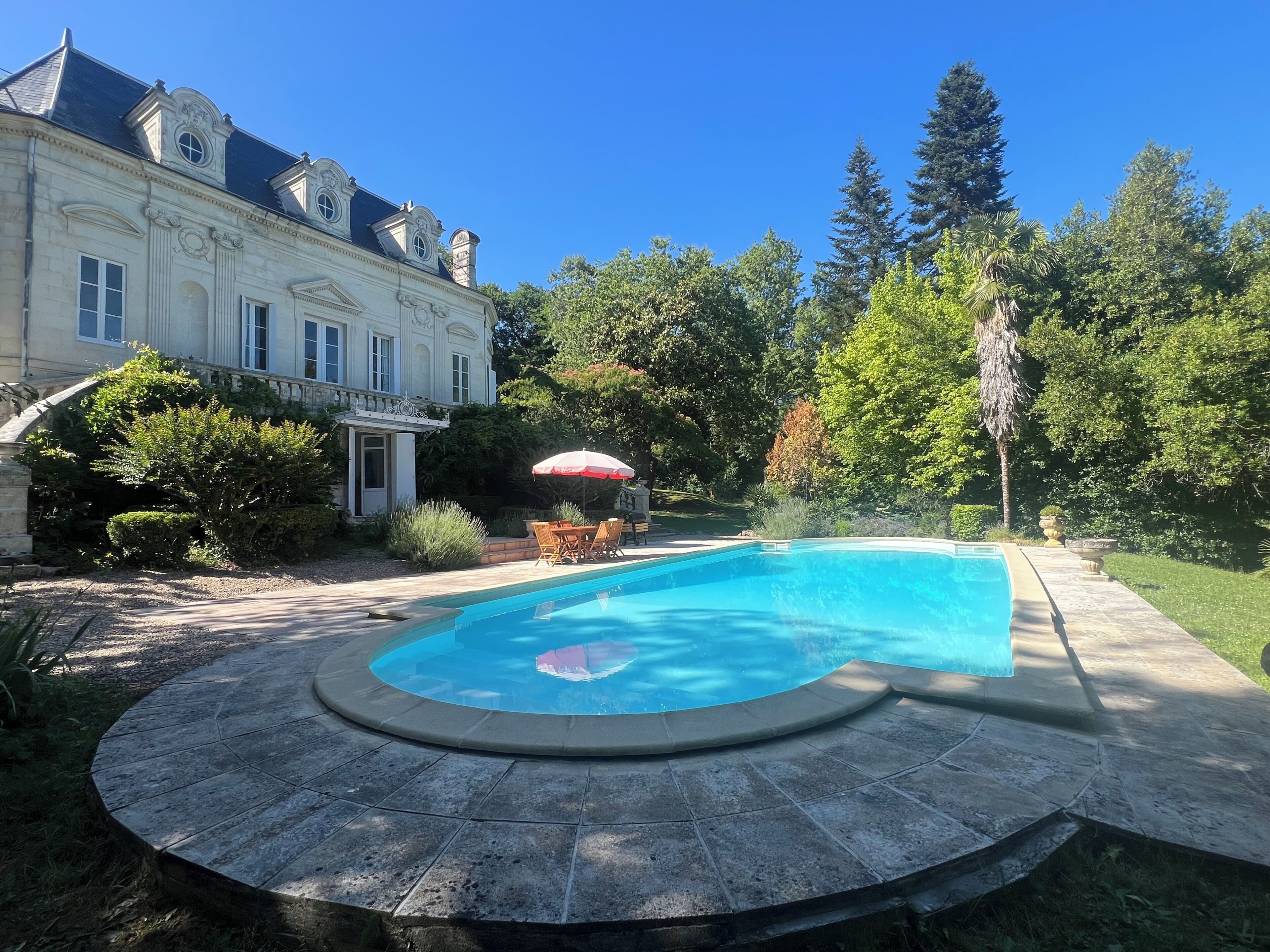 A swimming pool with a curved edge, a patio with outdoor furniture, and a large house with a balcony.
