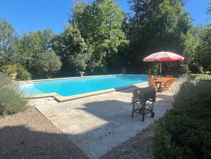 Ein Schwimmbad mit Terrassenbereich, umgeben von Bäumen und einem Grill.