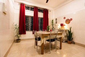 Een eetruimte met een tafel voor vier personen, rode gordijnen, potplanten en wanddecoraties.
