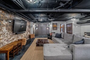 Un salon moderne avec un mur en briques, un grand téléviseur à écran plat, un canapé d’angle gris, une table basse en bois et un coin repas avec une table et des chaises en bois.