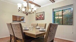 A dining room with a wooden table, four upholstered chairs, a chandelier, a painting of wine bottles, and a window with a view of trees. - 3BR Condo Near Ober Mountain & Downtown Gatlinburg (Gatlinburg)