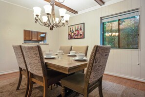 A dining room with a wooden table, four upholstered chairs, a chandelier, a painting of wine bottles, and a window with a view of trees. - 3BR Condo Near Ober Mountain & Downtown Gatlinburg (Gatlinburg)