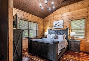 A wooden cabin bedroom with a bed, bedside tables, and a chandelier. - 4BR Smoky Mountain Cabin, Hot Tub & Theater (Sevierville)