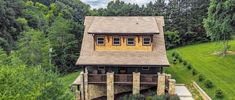 A two-story house with a stone foundation and a wooden upper level, surrounded by lush greenery and a well-maintained lawn.