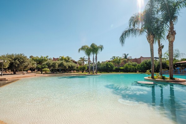 A clear swimming pool surrounded by palm trees and a building with a red roof.