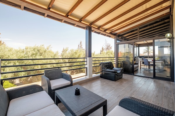 An outdoor patio with wicker furniture, a view of trees, and a glass door leading to an indoor area.