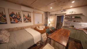 Una habitación acogedora con una cama, una mesa de madera, una silla, un escritorio, una lámpara y una cocina con un fregadero y armarios.
