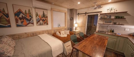 A cozy room with a bed, a wooden table, a chair, a desk, a lamp, and a kitchen area with a sink and cabinets.