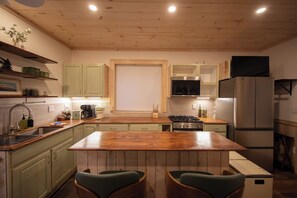 A kitchen with a wooden island, green cabinets, a stainless steel refrigerator, and a window with blinds.