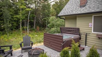 Een hottub met een houten bankje, een stoel en een kleine tafel in een tuin, met bomen en een huis op de achtergrond.