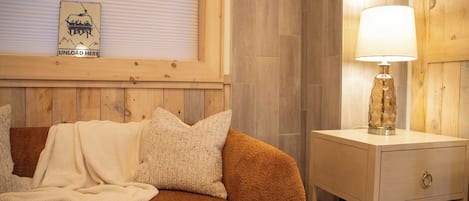 A cozy room with a brown sofa, a white lamp on a bedside table, and a window with blinds.