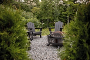 Un jardín con dos sillas Adirondack y un brasero de metal, rodeado de grava y follaje.