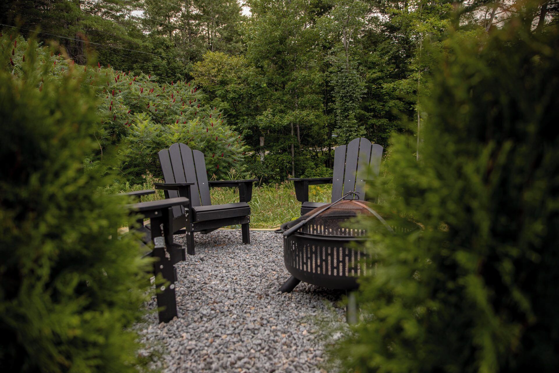 Un espace aménagé avec deux fauteuils Adirondack et un foyer en métal, le tout installé sur une surface de gravier et entouré d’une végétation luxuriante.