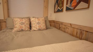 Una cama con colcha color beige y dos almohadones estampados, cabecera de madera y dos cuadros enmarcados en la pared.