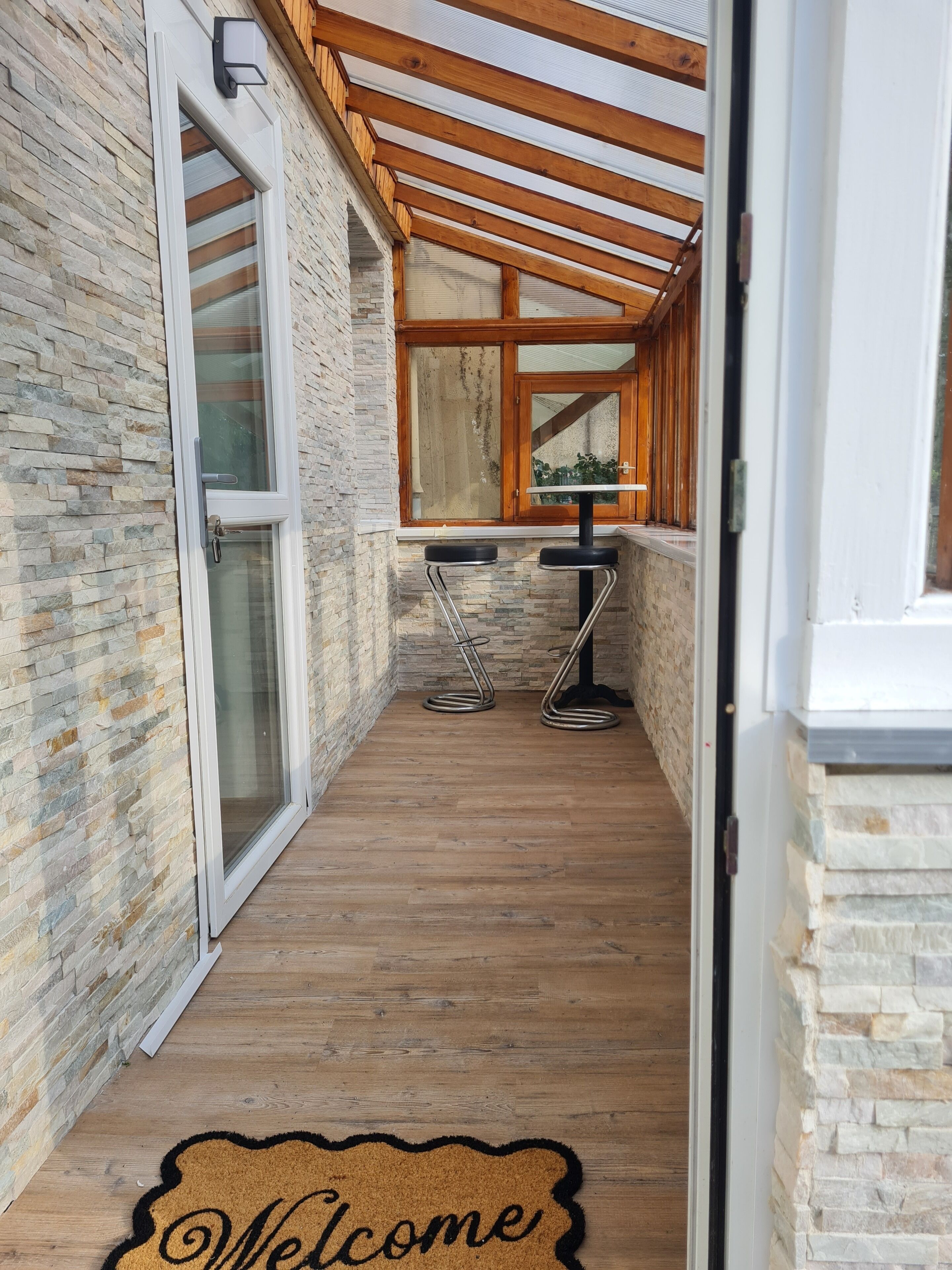 A corridor with a wooden floor, stone walls, and a glass door leading to a room. There are two metal bar stools and a window with wooden blinds.