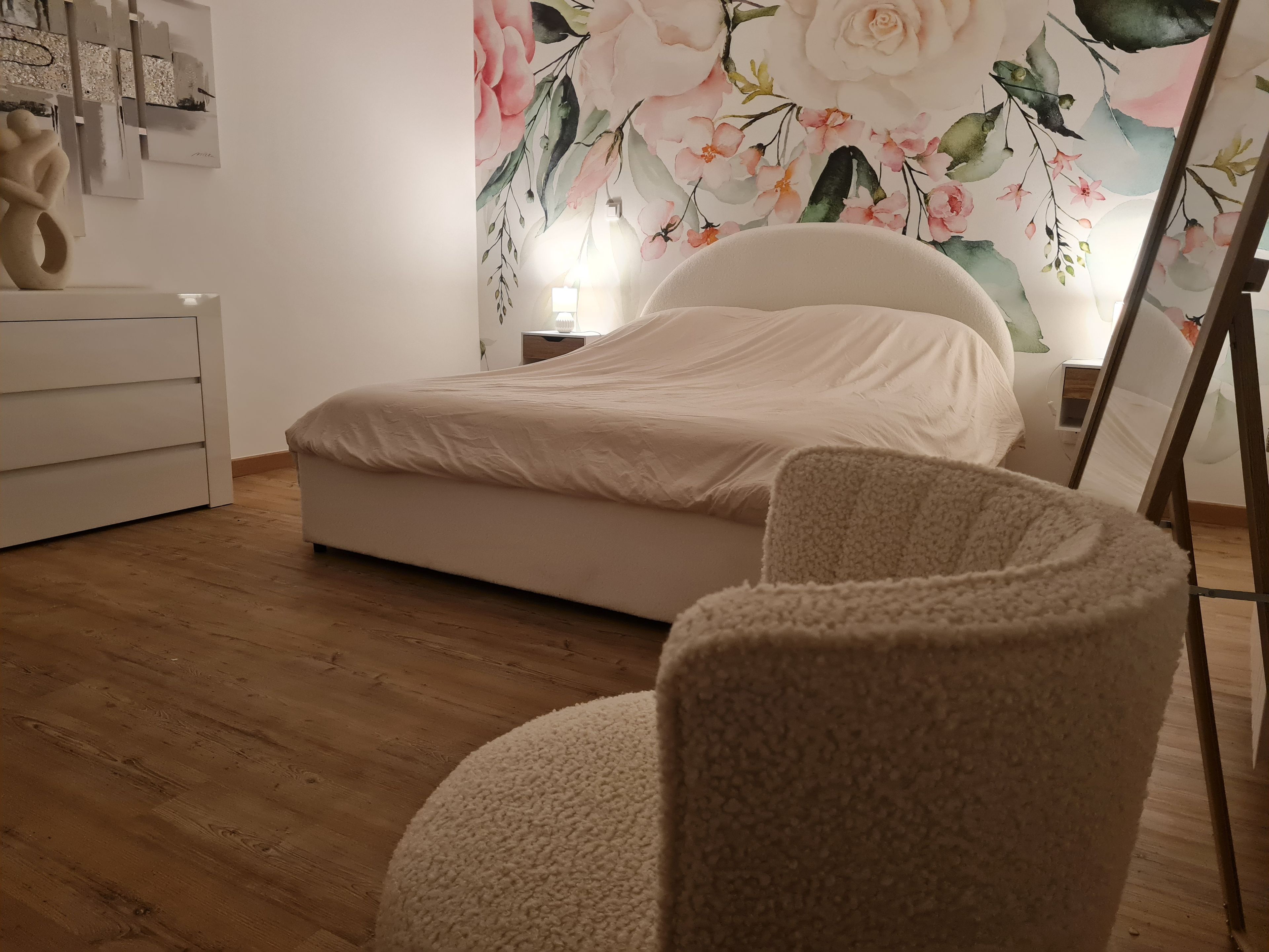 Une chambre à coucher avec un lit, une commode blanche, un fauteuil rond et une décoration murale à motifs floraux.
