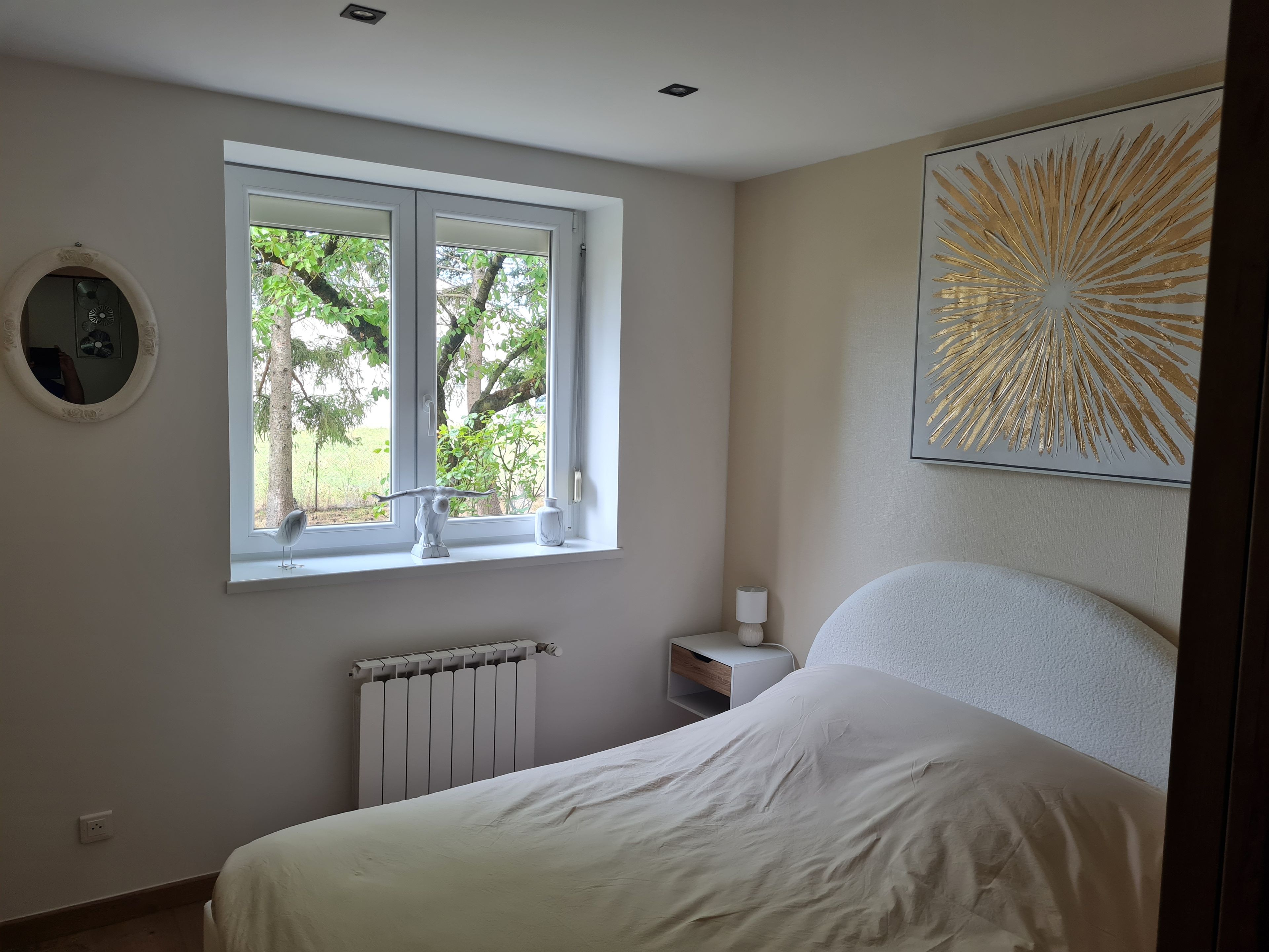 A bedroom with a bed, a window with a view of trees, a radiator, a bedside table with a lamp, and a wall-mounted mirror.