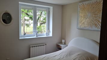 A bedroom with a bed, a window with a view of trees, a radiator, a bedside table with a lamp, and a wall-mounted mirror.