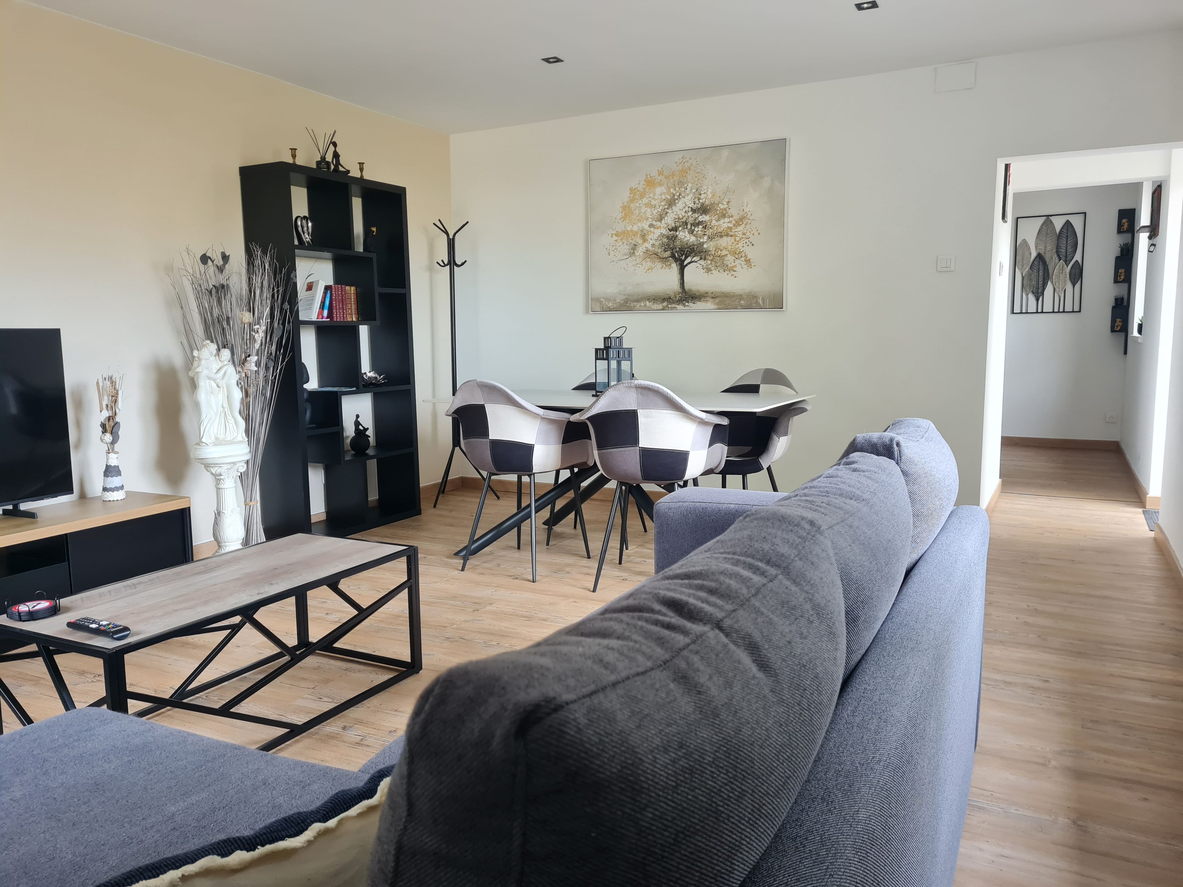 Un salon avec un canapé gris, un meuble de télévision noir, une table à manger entourée de chaises à carreaux et un tableau représentant un arbre.